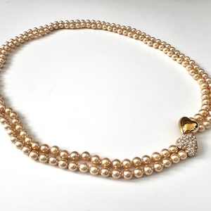 May include: A double-strand pearl necklace with a gold heart-shaped clasp. The clasp is adorned with clear crystals. The pearls are a light cream colour. The necklace is displayed on a white surface.