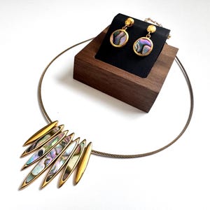 May include: A gold-toned necklace featuring a series of elongated, pointed pendants inlaid with iridescent abalone shell, paired with matching circular drop earrings. The jewelry set displays a metallic finish and natural shell patterns.