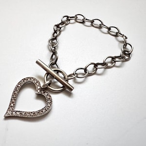 May include: A silver-toned chain bracelet featuring a toggle clasp and a heart-shaped pendant. The heart pendant is outlined with small, clear, sparkling stones along its entire perimeter, creating a bright and reflective appearance.