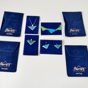 Könnte beinhalten: Eine Auswahl an geometrischem Schmuck in Blau und Grün, darunter ein Anhänger, ein Armband und Ohrringe, präsentiert auf dunkelblauem Samt. Jedes Schmuckstück wird auf einem passenden Samt-Display präsentiert, mit dem Markennamen "Biarritz" auf den Beuteln.
