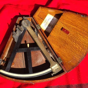 Può includere: Una scatola di legno con un coperchio incernierato, contenente un sestante in ottone e legno. Il sestante viene utilizzato per misurare gli angoli tra oggetti celesti, come il sole e l'orizzonte.
