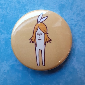 May include: A round, yellow button with a cartoon illustration of a figure with orange hair, white body, and two white feather-like shapes on top. The figure has a neutral expression. The button is glossy and has a slight reflection.
