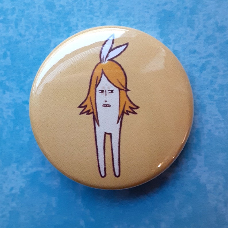 May include: A round, yellow button with a cartoon illustration of a figure with orange hair, white body, and two white feather-like shapes on top. The figure has a neutral expression. The button is glossy and has a slight reflection.