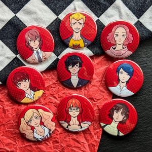 Op de afbeelding: Een verzameling van negen rode, ronde pins met illustraties van anime-personages. Elke pin heeft een ander karakterontwerp met verschillende haarkleuren en outfits. De pins zijn gerangschikt op een zwart-wit geruite stof.