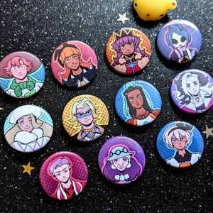 May include: A collection of round, colorful pinback buttons featuring various anime-style character illustrations. The buttons have a glossy finish and are arranged on a black, speckled surface with star-shaped confetti.