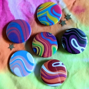 Puede incluir: Un conjunto de siete botones redondos con diseños coloridos y arremolinados. Los botones presentan varias combinaciones de colores, incluyendo azul, rosa, amarillo, morado y naranja. Varias pequeñas estrellas plateadas decoran la escena.