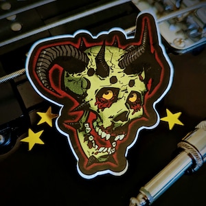 May include: A sticker featuring a green, yellow, and red demonic skull with horns. The sticker has a black border and is surrounded by yellow star-shaped stickers and a silver audio jack.