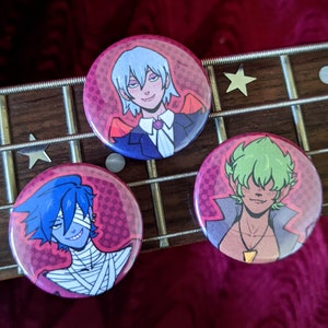 Op de afbeelding: Drie ronde buttons met illustraties van anime-personages. Elke button toont een ander personage met verschillend haar en kleding, tegen een roze achtergrond met een stippenpatroon. De buttons liggen op een houten oppervlak.