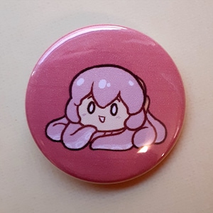 May include: A round, pink button featuring a cartoon character with pink hair and a smiling face. The character is drawn with black outlines and white highlights, set against a solid pink background. The button has a glossy finish.