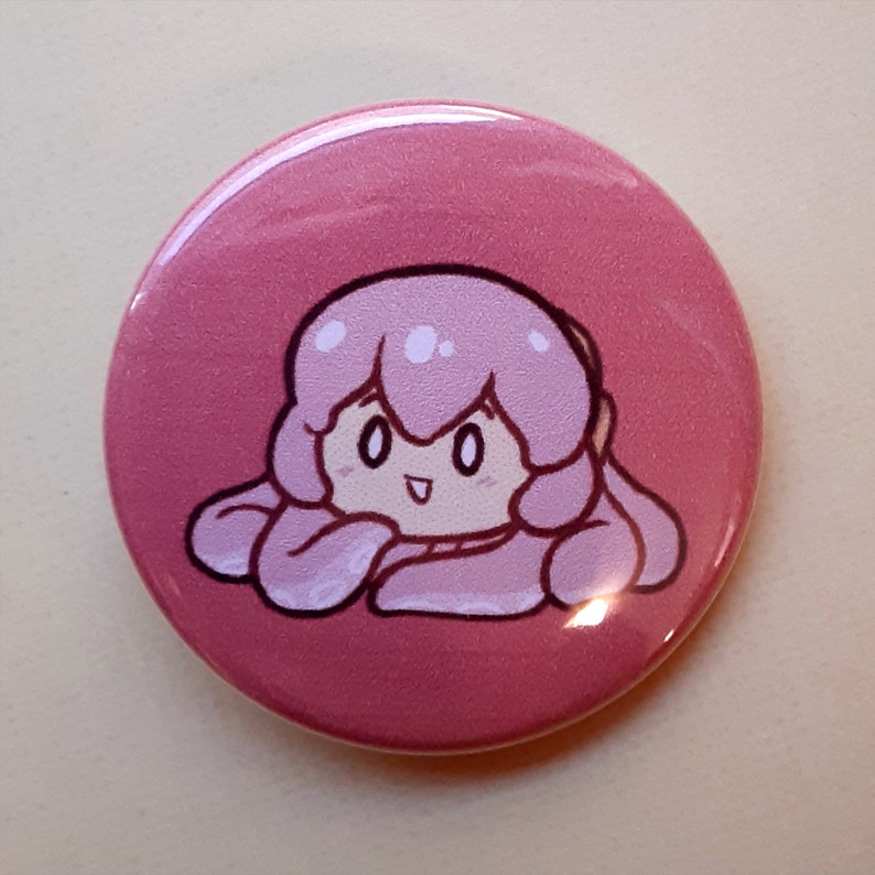 May include: A round, pink button featuring a cartoon character with pink hair and a smiling face. The character is drawn with black outlines and white highlights, set against a solid pink background. The button has a glossy finish.