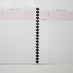 May include: Open notebook with a black spiral binding, featuring a "Badge Tracker" title. The pages are white with a grid pattern and pink-colored header sections for "Scout Name" and "Badge/Journey". The notebook is open on a white surface.