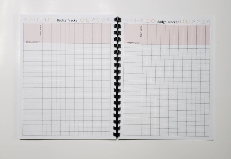 May include: Open notebook with a black spiral binding, featuring a "Badge Tracker" title. The pages are white with a grid pattern and pink-colored header sections for "Scout Name" and "Badge/Journey". The notebook is open on a white surface.