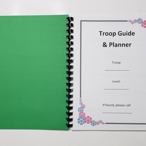 May include: Open spiral-bound notebook with a green cover and a white page with the text "Troop Guide & Planner." The page includes fill-in-the-blank lines for troop and level, and a contact request. Floral corner accents.