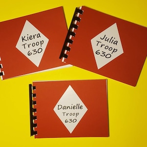 Puede incluir: Tres cuadernos rojos de espiral con etiquetas blancas en forma de diamante. Las etiquetas tienen texto negro que dice "Kiera Troop 630", "Julia Troop 630" y "Danielle Troop 630".