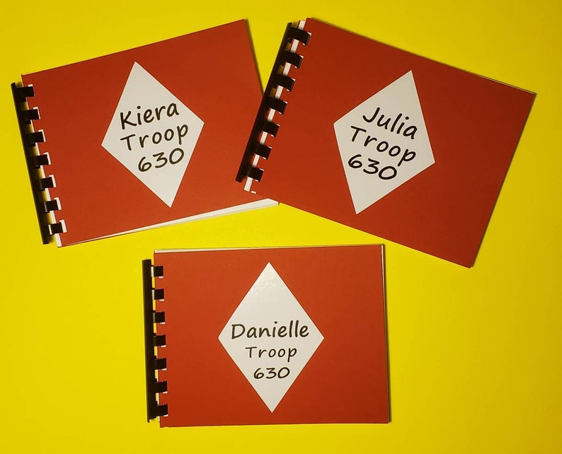 Puede incluir: Tres cuadernos rojos de espiral con etiquetas blancas en forma de diamante. Las etiquetas tienen texto negro que dice "Kiera Troop 630", "Julia Troop 630" y "Danielle Troop 630".
