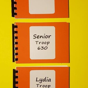 Puede incluir: Tres cuadernos naranjas con etiquetas blancas. El cuaderno superior dice "Senior". El cuaderno del medio dice "Senior Troop 630". El cuaderno inferior dice "Lydia Troop 630".