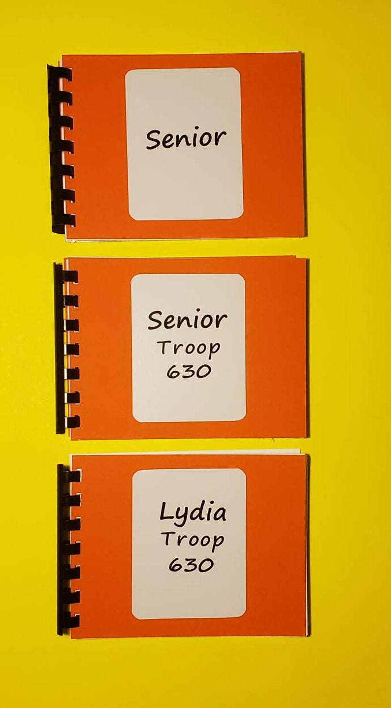Puede incluir: Tres cuadernos naranjas con etiquetas blancas. El cuaderno superior dice "Senior". El cuaderno del medio dice "Senior Troop 630". El cuaderno inferior dice "Lydia Troop 630".