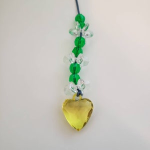May include: A yellow heart-shaped crystal charm hangs from a string of green and clear glass beads. The beads are shaped like flowers.