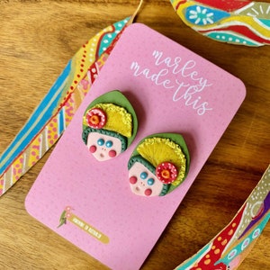 May include: A pair of handmade polymer clay earrings featuring a whimsical design with green, yellow, pink, and red details. The earrings are displayed on a pink card with the text "marley made this" and "handmade in australia".
