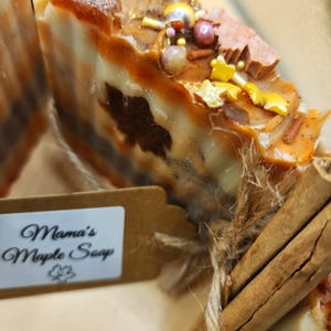 Könnte beinhalten: Eine handgemachte Seife in Form eines Blocks mit einem braunen und weißen Wirbelmuster, bedeckt mit Streuseln und Goldflocken. Die Seife ist mit Schnur und Zimtstangen gebunden. Das Etikett lautet "Mama's Maple Soap".