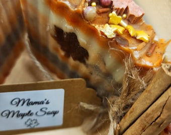 May include: A bar of handmade soap with a brown and white swirl design, topped with sprinkles and gold flakes. The soap is tied with twine and cinnamon sticks. The label reads "Mama's Maple Soap".