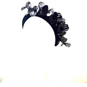 May include: A black floral crown with a crescent-shaped base. The crown features multiple black and silver-toned flowers of varying sizes. The flowers are arranged along the top of the crown, creating a decorative headpiece.