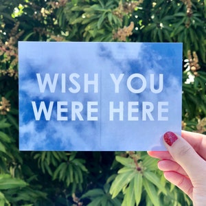 May include: A blue and white postcard with the text "Wish You Were Here" against a cloudy sky background.