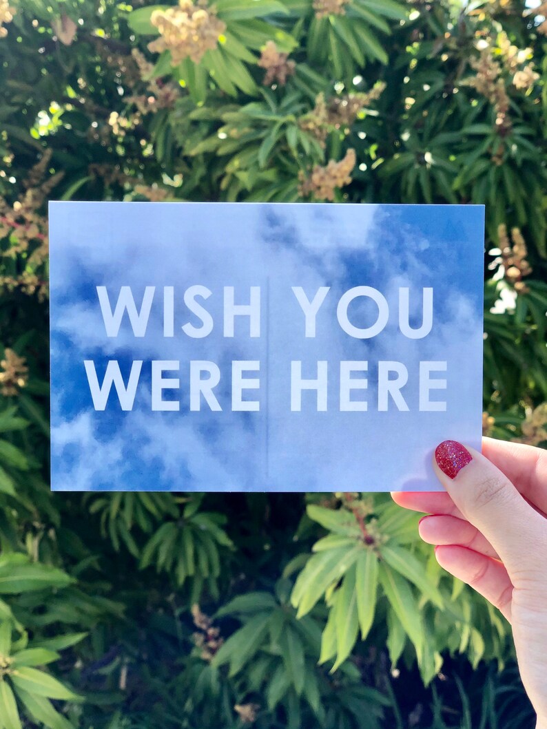 May include: A blue and white postcard with the text "Wish You Were Here" against a cloudy sky background.