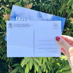 May include: A white postcard with a blue sky and clouds design. The front of the postcard has the text "Luna Peak" and "Serving the cancer and grief communities - celebrate life with us! www.lunapeakfoundation.org". The back of the postcard has a space for a stamp and an address.