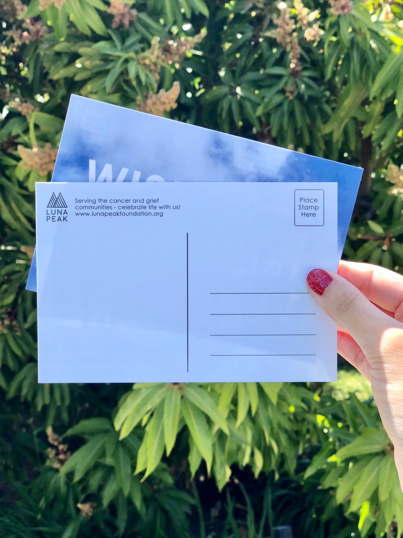 May include: A white postcard with a blue sky and clouds design. The front of the postcard has the text "Luna Peak" and "Serving the cancer and grief communities - celebrate life with us! www.lunapeakfoundation.org". The back of the postcard has a space for a stamp and an address.