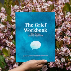 May include: A blue book with the title "The Grief Workbook" and the subtitle "Grief Tested, Mourner Approved!"  The book has a speech bubble with the text "I miss you" in it. The book is being held by a hand in front of a background of pink flowers.
