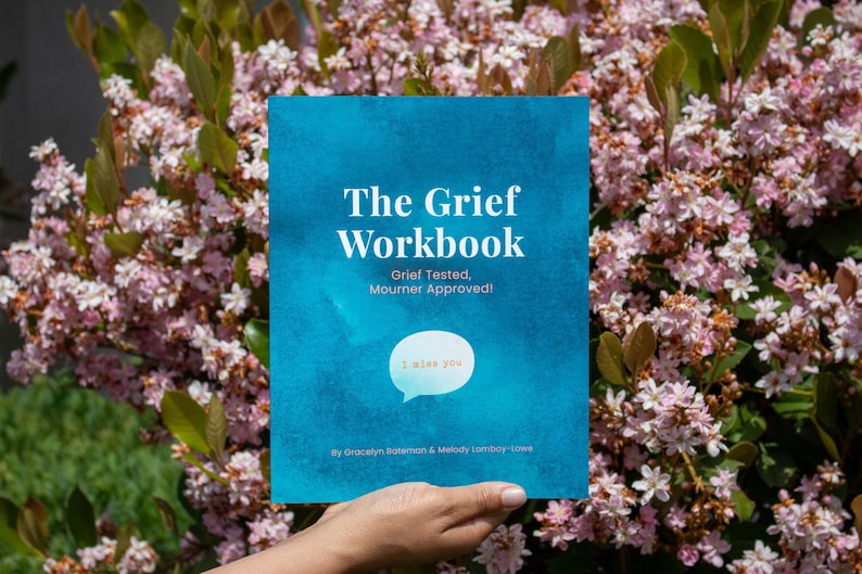 May include: A blue book with the title "The Grief Workbook" and the subtitle "Grief Tested, Mourner Approved!"  The book has a speech bubble with the text "I miss you" in it. The book is being held by a hand in front of a background of pink flowers.