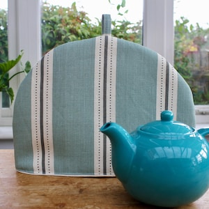 May include: A teal ceramic teapot with a matching lid and handle stands in front of a teal and white striped tea cosy. The tea cosy has a handle at the top. The background includes a window and plants.