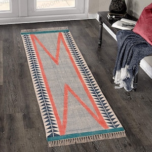 May include: A long, rectangular rug with a geometric pattern. The rug has a white background with a red and blue design. The rug has a fringe border.