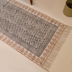 May include: A white cotton runner rug with a black and brown geometric pattern. The rug has a fringe border and is perfect for adding a touch of style to any room.