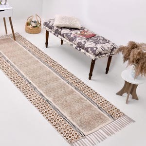 Puede incluir: Una alfombra de pasillo beige y marrón estampada con flecos. La alfombra tiene un campo central beige bordeado por diseños geométricos y florales en marrón y azul marino. La alfombra está sobre un suelo blanco.
