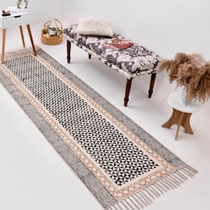 Op de afbeelding: Een lange, rechthoekige vloerkleed met een geometrisch zwart-wit patroon. Het vloerkleed heeft een beige rand met een decoratief ontwerp en franjes aan de uiteinden. Het vloerkleed ligt op een witte vloer.