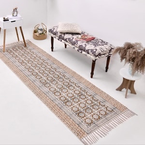 May include: A long, rectangular rug with a repeating floral and geometric pattern in brown, beige, and white. The rug has a fringed edge and is placed on a white floor. The rug is part of a home decor theme.