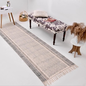 Puede incluir: Una alfombra de pasillo color crema con un patrón geométrico y bordes con flecos. La alfombra presenta un diseño repetitivo de diamantes y puntos en negro y gris, bordeado por una franja negra y gris. La alfombra está colocada sobre un suelo blanco.