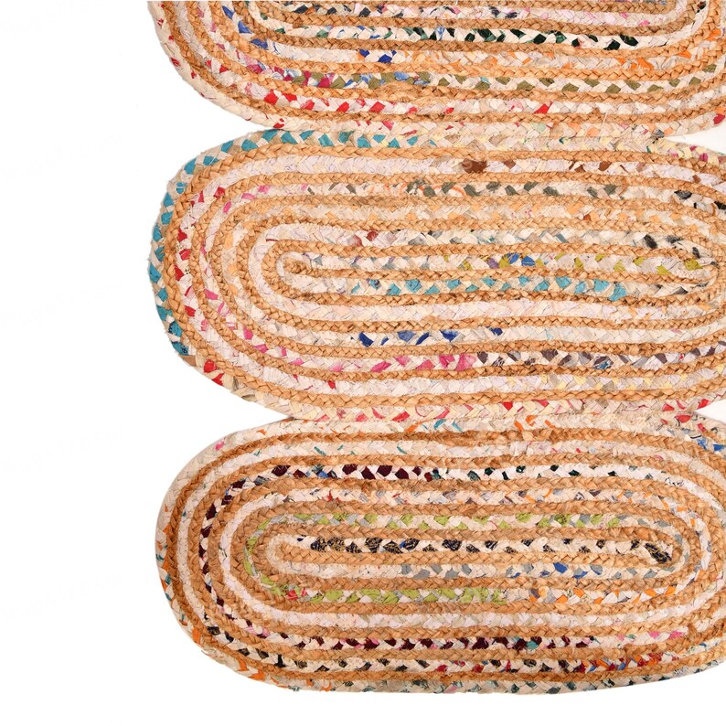May include: Close-up of three oval-shaped jute rugs. The rugs have a natural tan color with a braided border. The centers are filled with colorful recycled fabric scraps, creating a textured, multi-colored pattern. The rugs are arranged vertically.