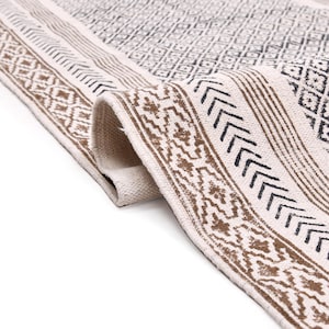 Könnte beinhalten: Eine aufgerollte, cremefarbene Decke mit geometrischem Muster. Die Decke hat ein sich wiederholendes Rautenmuster in der Mitte, das von braunen und schwarzen Zierbändern mit verschiedenen geometrischen Designs umrandet wird. Die Decke besteht aus einem gewebten Material.