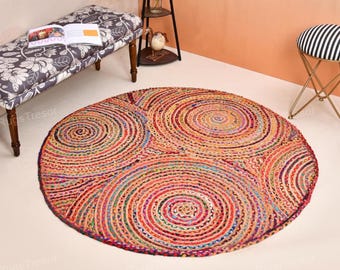 Op de afbeelding: Een ronde, veelkleurige jute vloerkleed met een spiraalvormig ontwerp. Het vloerkleed heeft concentrische cirkels in tinten rood, oranje, blauw en paars. Een bank met bloemenpatroon, een kleine kruk en een decoratief rek zijn ook zichtbaar.