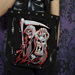 Puede incluir: Bolso tote negro con un diseño bordado de una parca y una calavera. El bolso tiene tachuelas plateadas en el borde superior y detalles de imperdibles. El bolso se sujeta con correas negras.