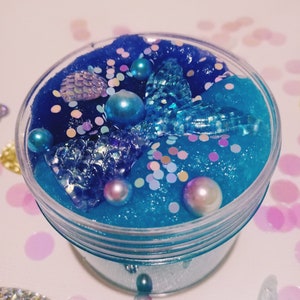 K&ouml;nnte beinhalten: Ein klares Glas gef&uuml;llt mit blauem Schleim mit schimmerndem Glitzer, Perlen und einem blauen Meerjungfrauen-Schwanz-Anh&auml;nger.