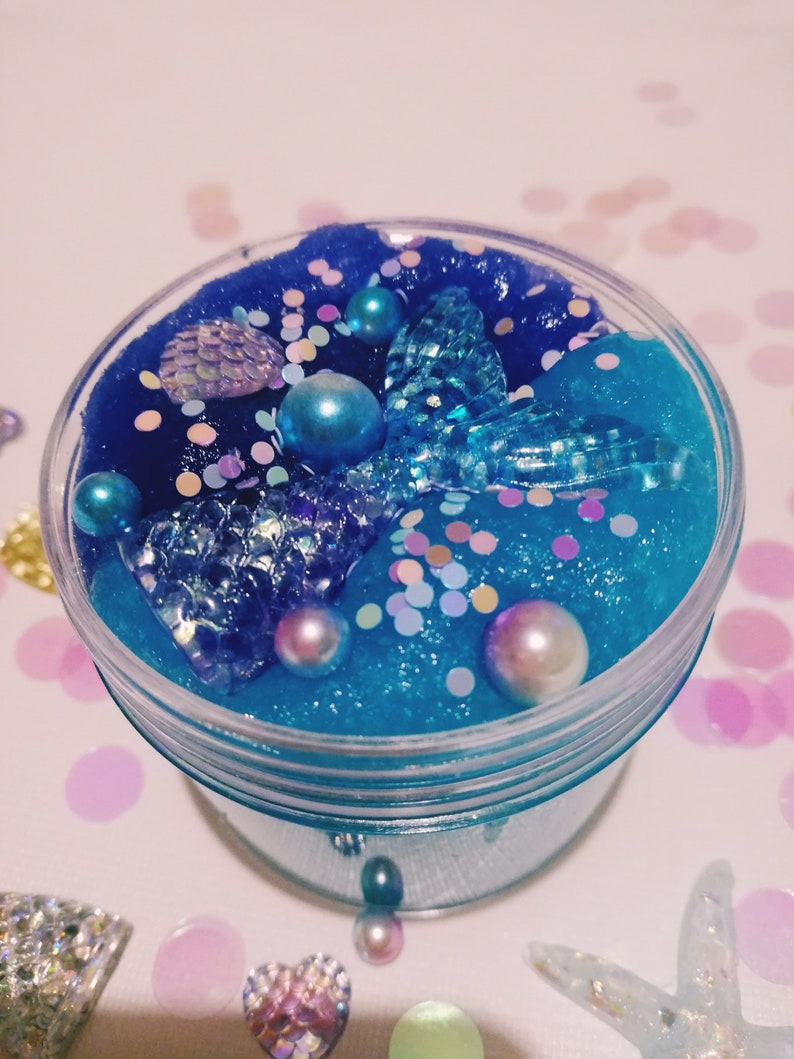 K&ouml;nnte beinhalten: Ein klares Glas gef&uuml;llt mit blauem Schleim mit schimmerndem Glitzer, Perlen und einem blauen Meerjungfrauen-Schwanz-Anh&auml;nger.