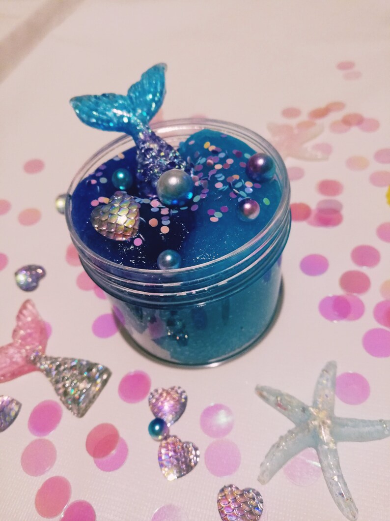 K&ouml;nnte beinhalten: Ein blauer Schleim mit Glitzer und schillernden Perlen in einem durchsichtigen Glas mit einer Meerjungfrauenflosse oben drauf. Das Glas ist von rosa und wei&szlig;en Konfetti umgeben.