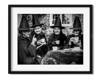 May include: Four women wearing black hats and cloaks are sitting at a table, drinking tea and talking. The women are all smiling and seem to be enjoying each other's company.