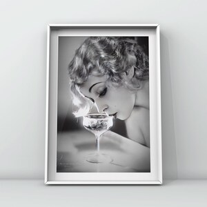 Op de afbeelding: Een zwart-witfoto van een vrouw met kort, golvend haar die aan een glas champagne ruikt.