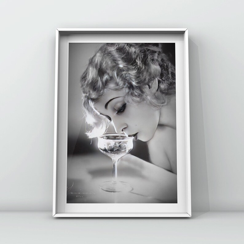 Op de afbeelding: Een zwart-witfoto van een vrouw met kort, golvend haar die aan een glas champagne ruikt.