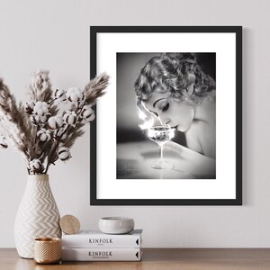 Op de afbeelding: Zwart-witfoto van een vrouw in een vintage jurk, die naar een glas champagne kijkt. De afbeelding is ingelijst in een zwart frame met een witte passe-partout.